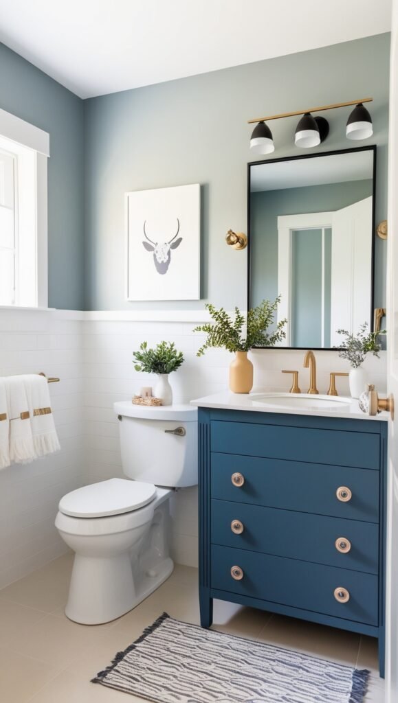 Small modern bathroom with modern decor and dark blue vanity with gold handles and black gold lights hanging above a large mirror with black trim gold and white towels hanging on gold shower towel bar and small throw rug in front of the sink 